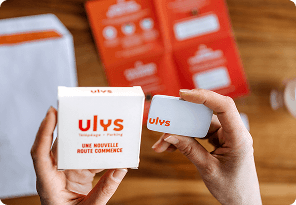 Personne tenant un badge télépéage et sa boîte au‑dessus d’une table avec des documents et supports posés en arrière-plan.